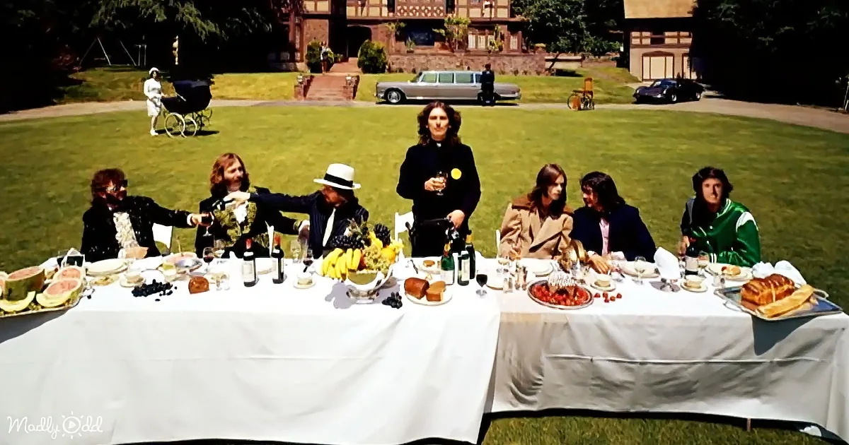 A man stands at a long table with six seated individuals, surrounded by food and drinks, in a grassy yard with a house...