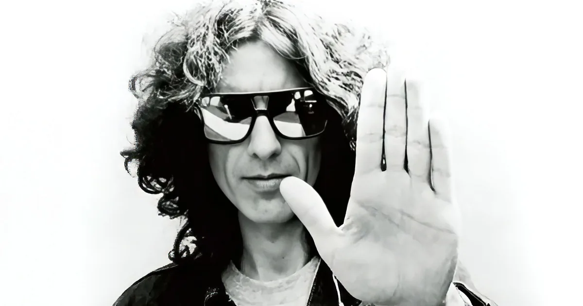A person with long, curly hair and sunglasses holds up a hand with fingers extended.