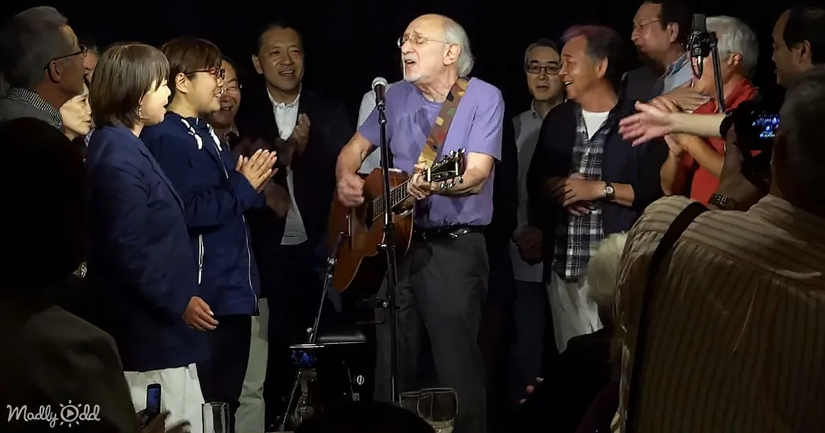 A man in a purple shirt plays guitar and sings in front of a group of clapping people.