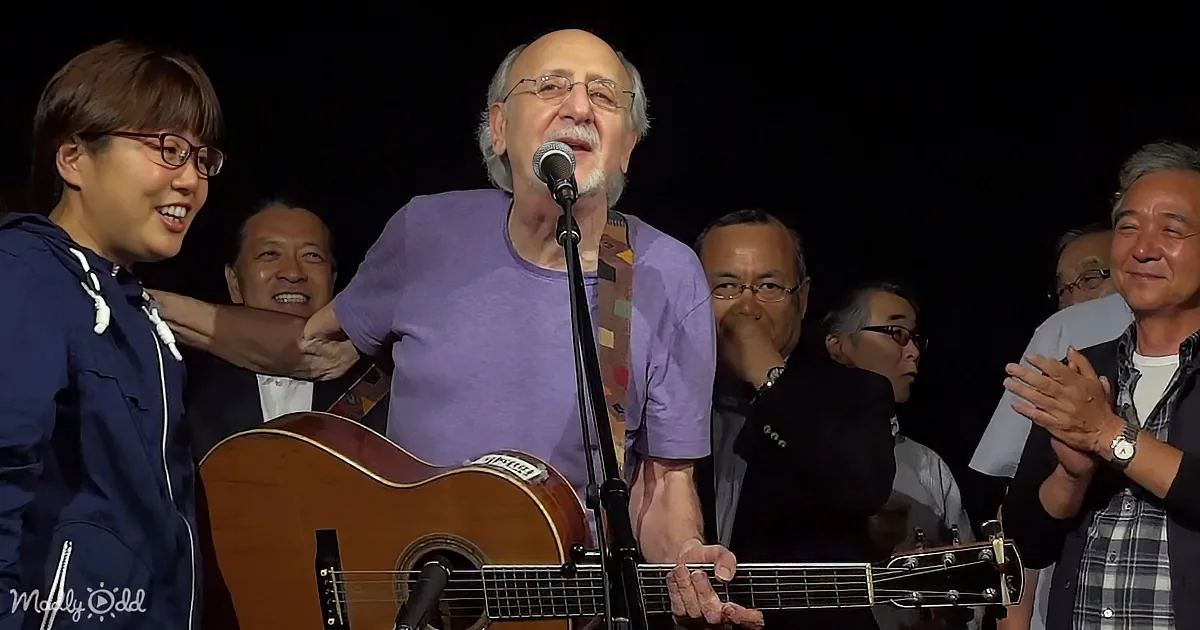 A man in a purple shirt sings into a microphone while holding an acoustic guitar, surrounded by smiling people.