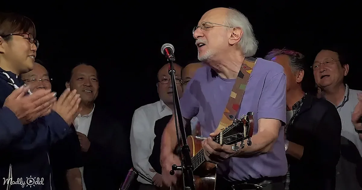 An older man plays guitar and sings into a microphone, surrounded by clapping people.