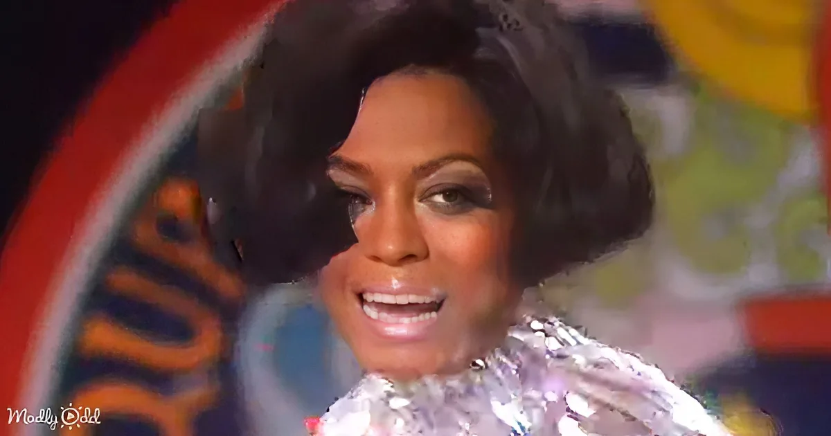 A person with a large, curly hairstyle and a shiny, reflective outfit is smiling.