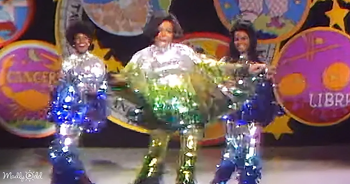 Three performers in shiny, reflective costumes stand on stage with colorful, whimsical background.