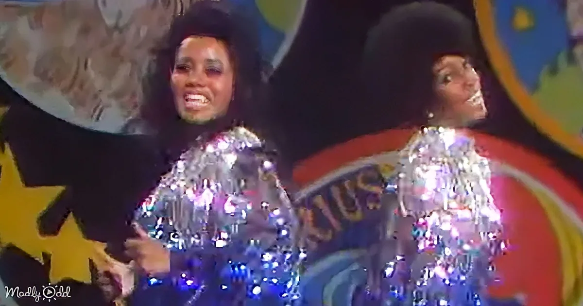 Two women in sparkling silver outfits perform on stage, smiling and dancing.