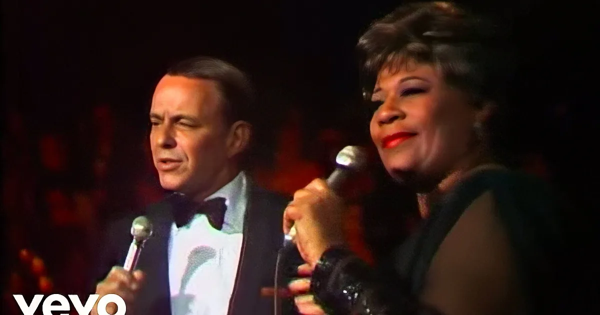 A man in a suit and a woman in a black dress hold microphones, possibly performing on stage.