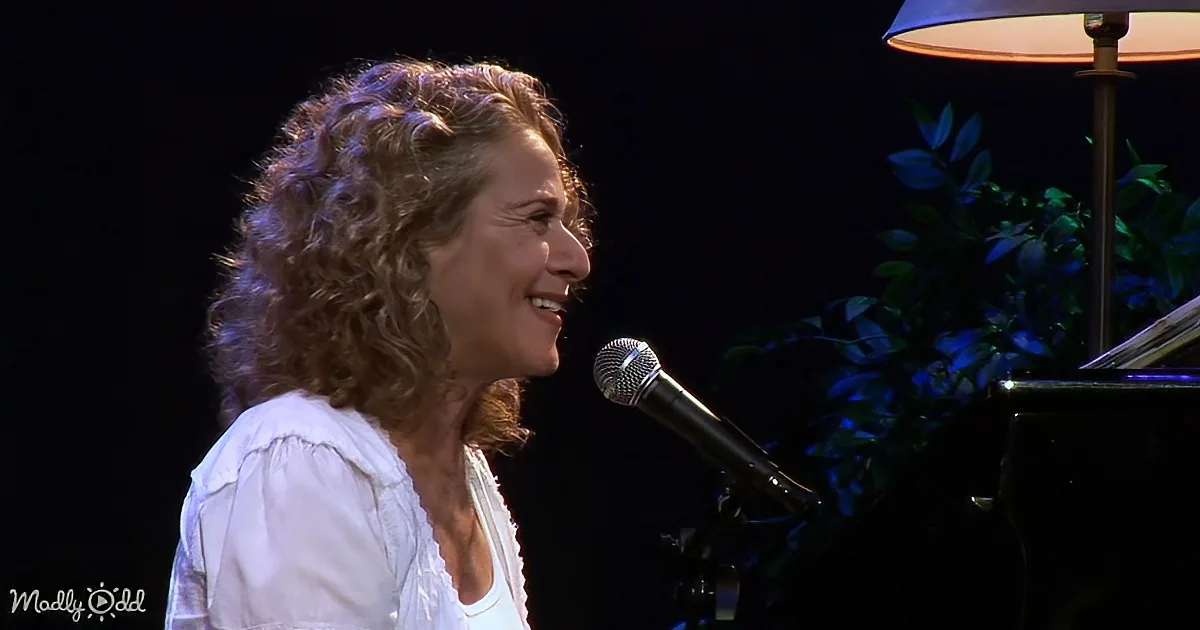 A woman with curly hair sings into a microphone, wearing a white shirt, with a piano and plant in the background.