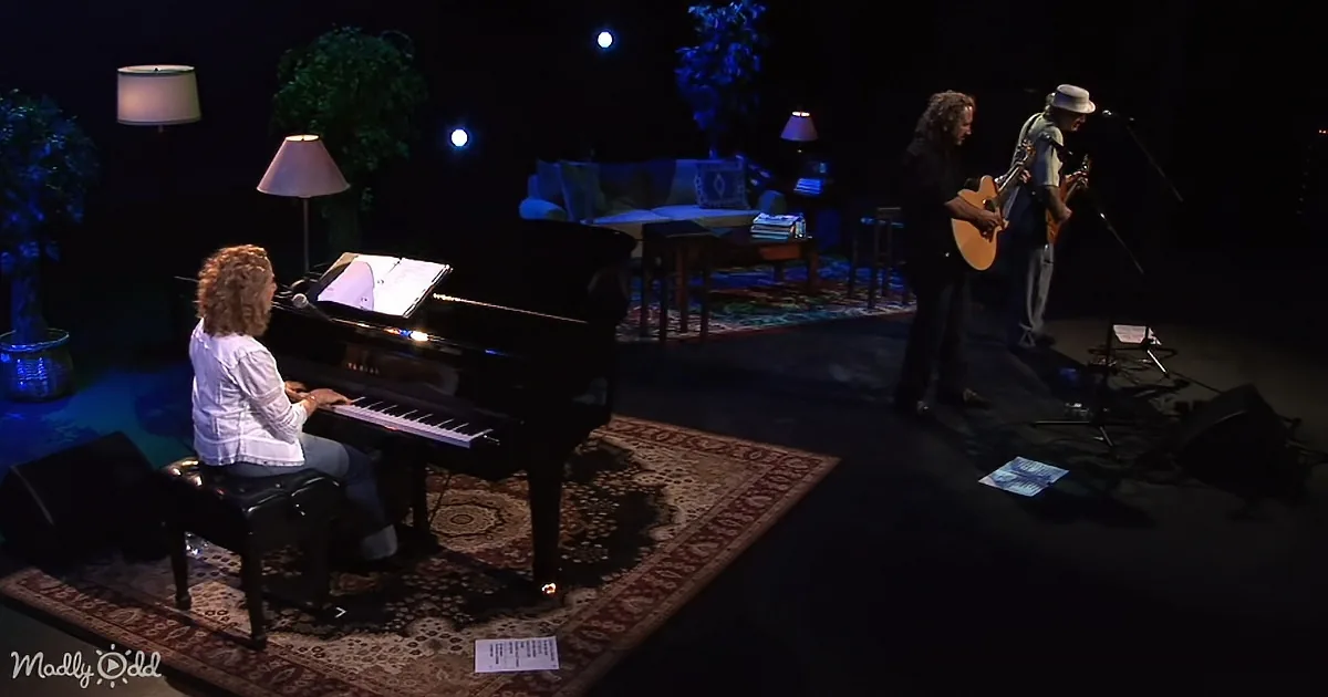 A woman plays piano while another person sings and plays guitar on a dimly lit stage.