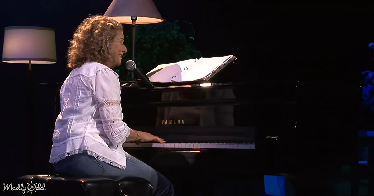 A woman in a white blouse sits at a piano, singing into a microphone with sheet music in front of her.