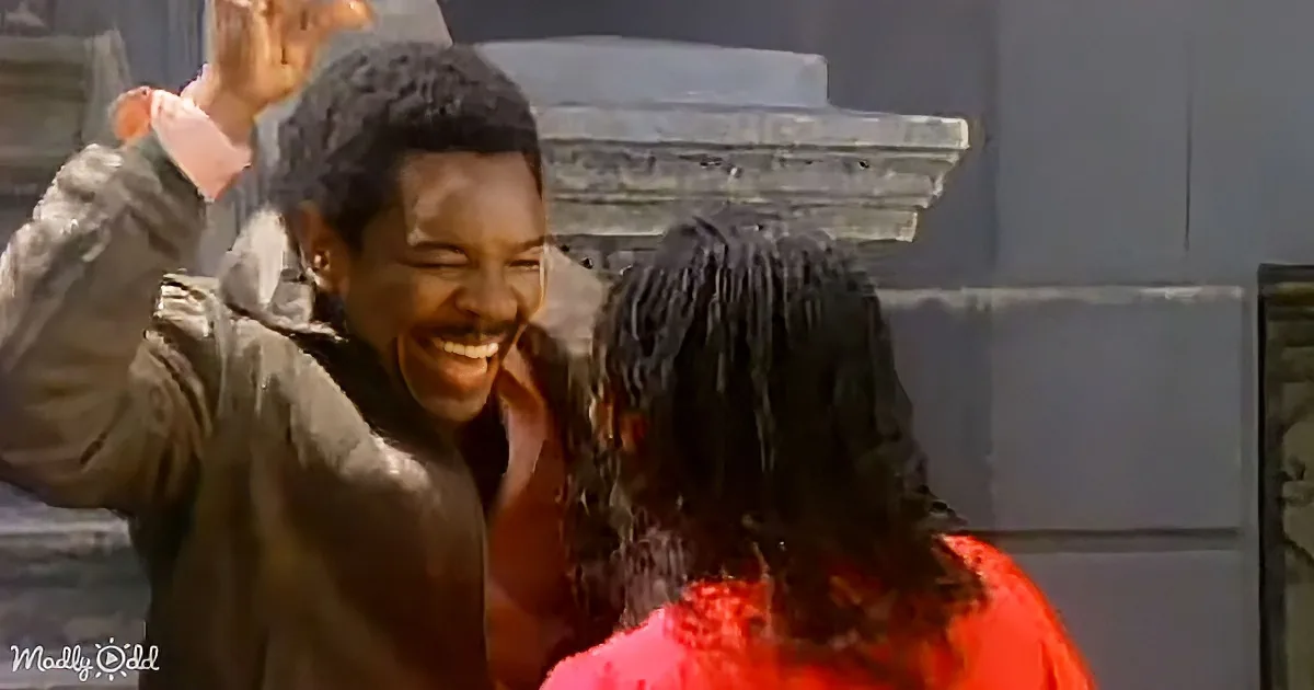 A man in a brown jacket smiles widely, raising his hand, while a woman in a red outfit stands beside him.