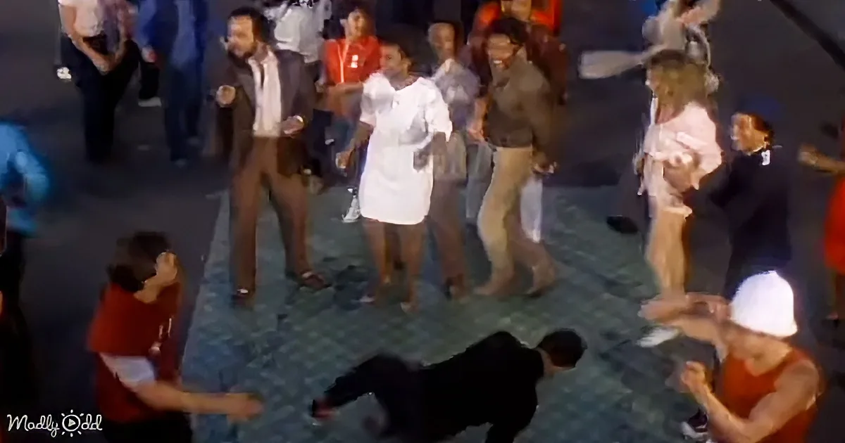 A man in a suit is dancing with a woman in a white dress, surrounded by a crowd of people.