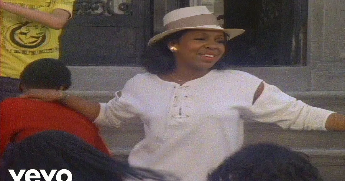 A smiling woman in a white shirt and hat sits on steps, surrounded by people.