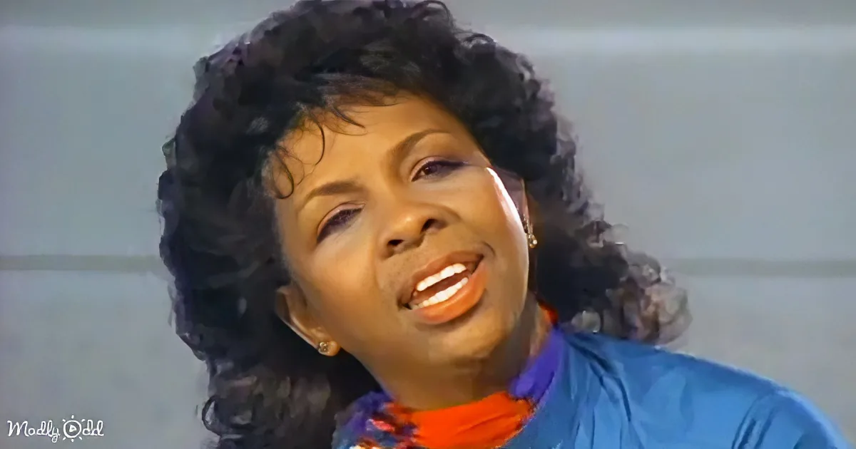 A woman with curly hair, wearing a blue jacket and a colorful scarf, is singing with her eyes closed.