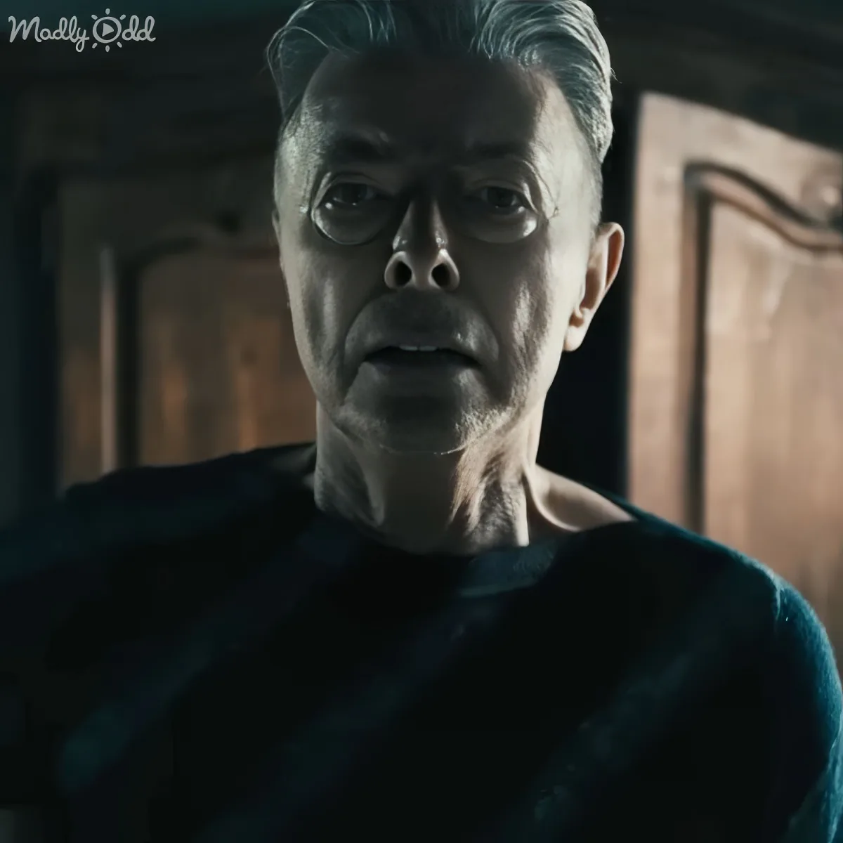 An older man with gray hair, wearing a dark shirt, stands in a dimly lit room with wooden doors.