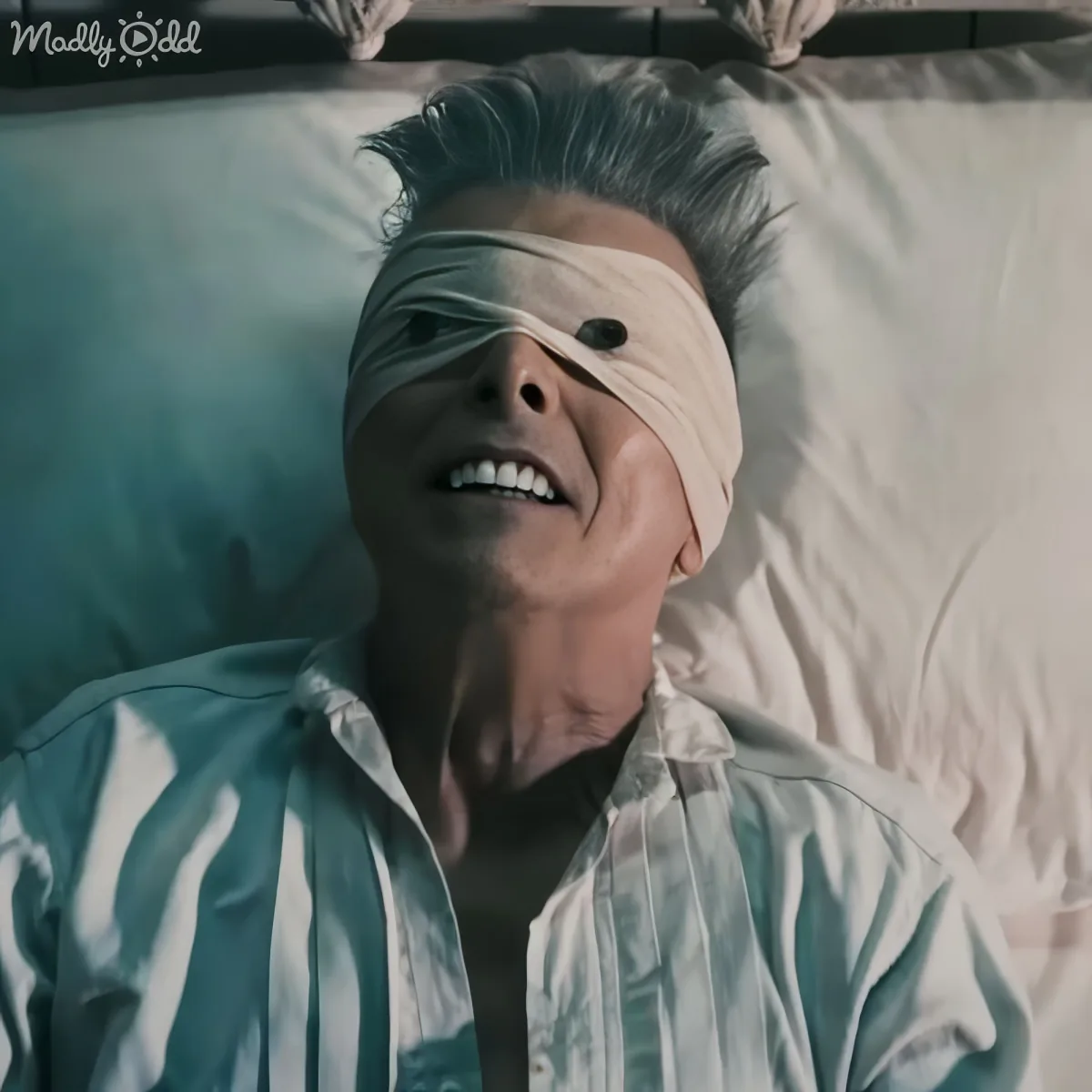 A person in a striped shirt lies in bed with a white cloth covering their eyes, mouth slightly open.