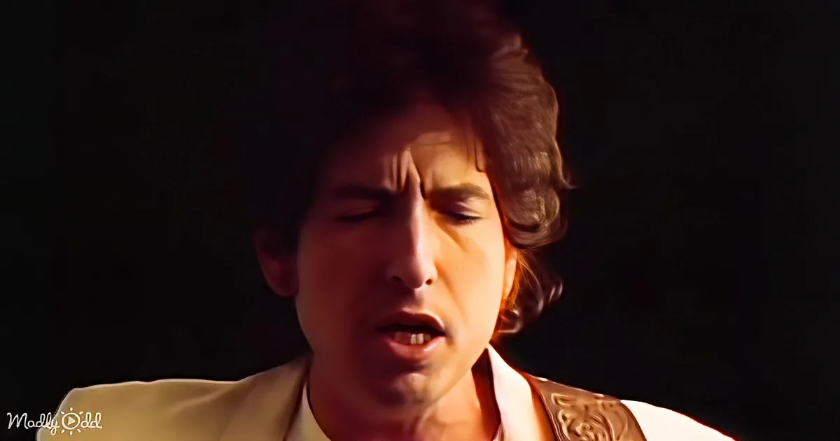 A person with curly hair and a light-colored suit is singing with their eyes closed.