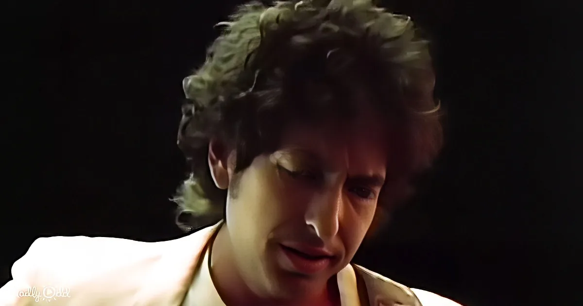 A person with curly hair and a light-colored shirt is shown in a close-up, looking downward with a dark background.