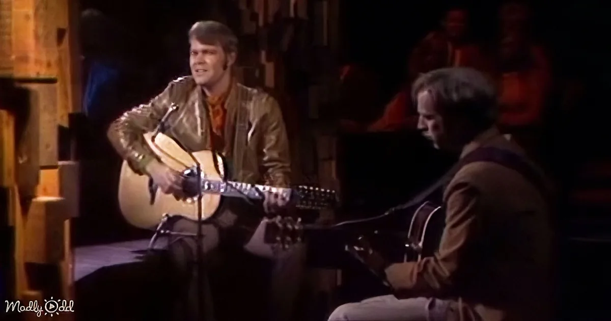 A man in a leather jacket plays an acoustic guitar on stage, accompanied by another musician.