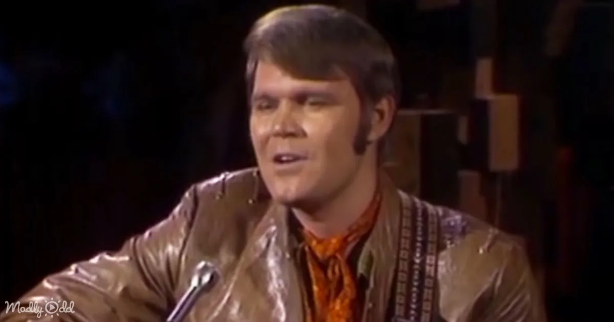 A man in a shiny brown jacket and orange shirt is speaking into a microphone.