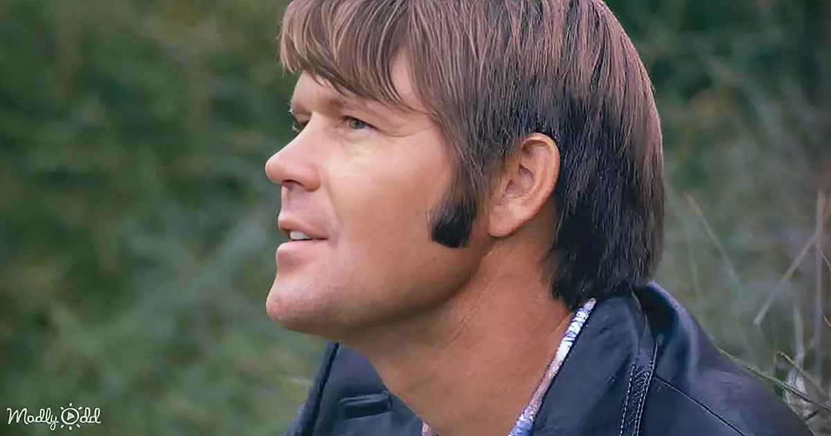 A man with long hair and a leather jacket looks upward, set against a blurred green background.