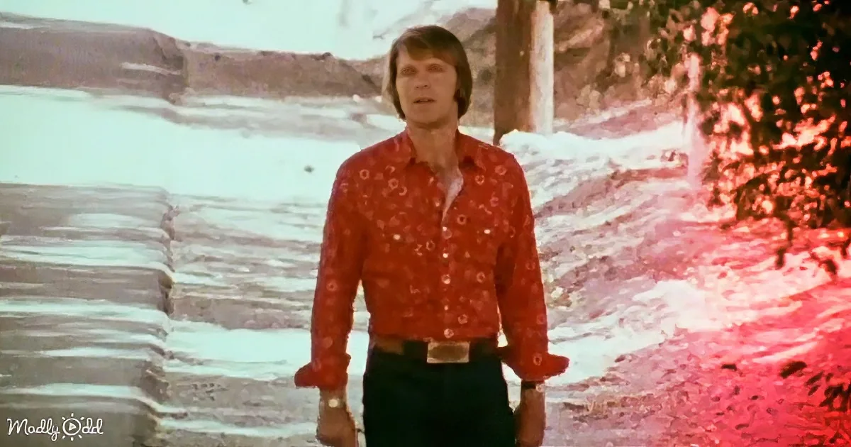 A man in a red patterned shirt stands outdoors near water, with a scenic background.
