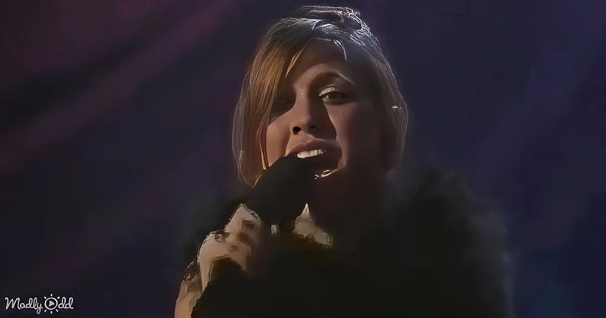 A person singing into a microphone, wearing a black outfit, with a dark background and some light reflections.