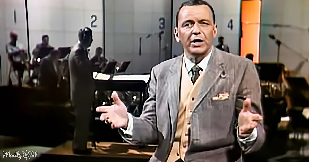 A man in a suit gestures with his hands, standing in front of a band and numbered doors.