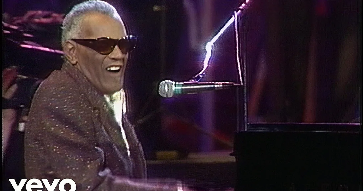 A man in sunglasses and a suit sings into a microphone while playing a piano.