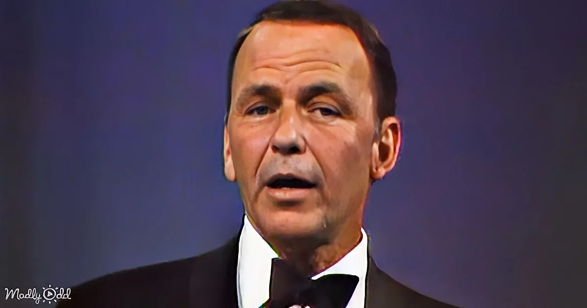 A man in a suit and bow tie speaks into a microphone against a dark background.