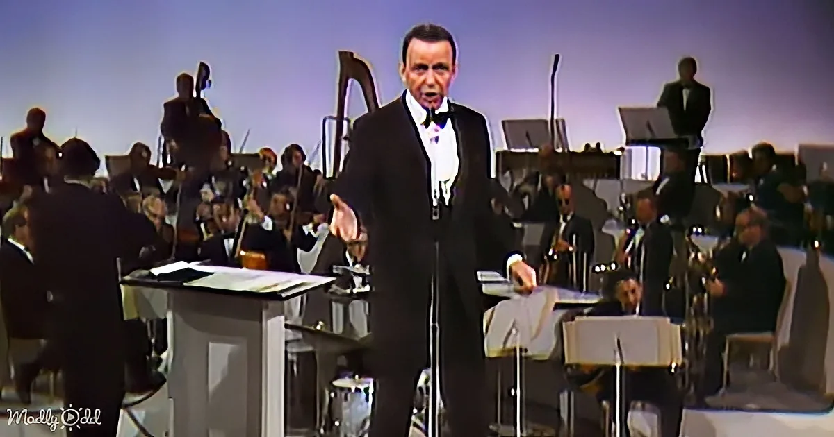 A man in a tuxedo sings passionately in front of an orchestra.