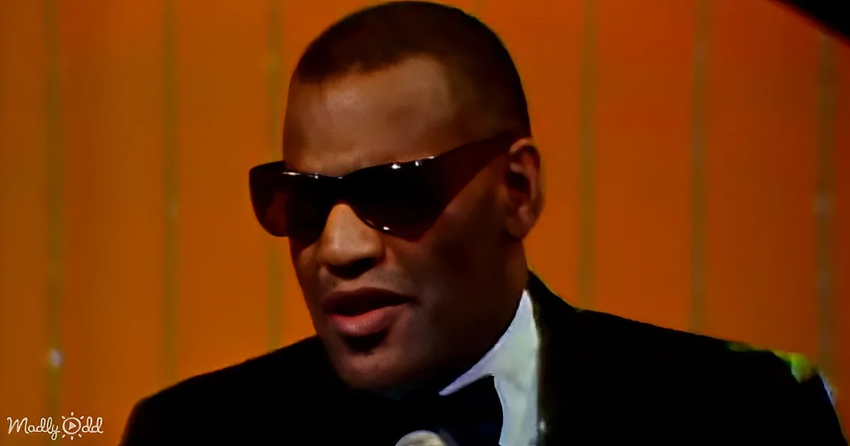 A man in a suit and sunglasses is speaking into a microphone, with an orange background.