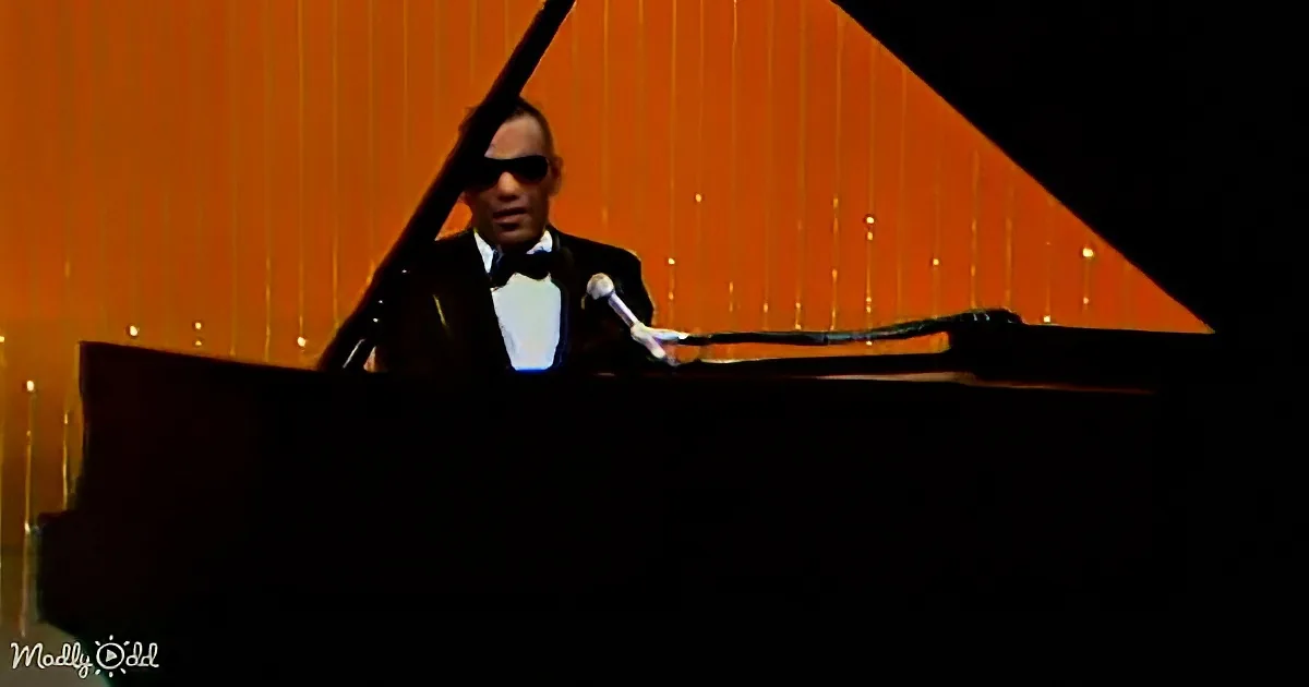A man in a tuxedo and sunglasses plays a grand piano on a stage with orange lighting.