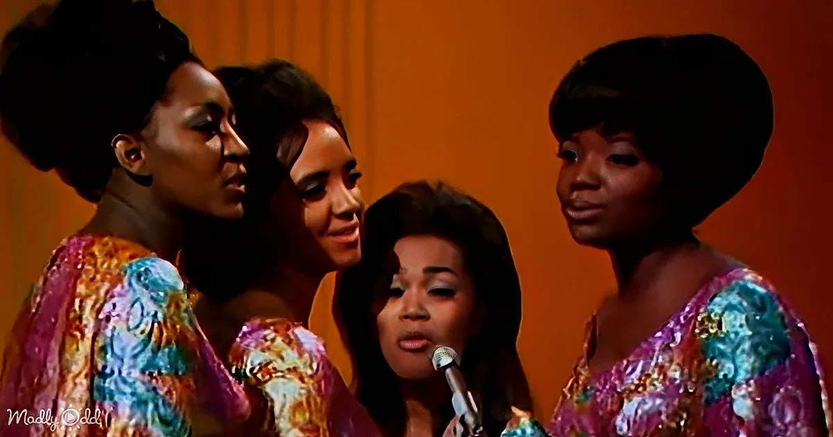A woman sings into a microphone, surrounded by three women in colorful, patterned dresses.