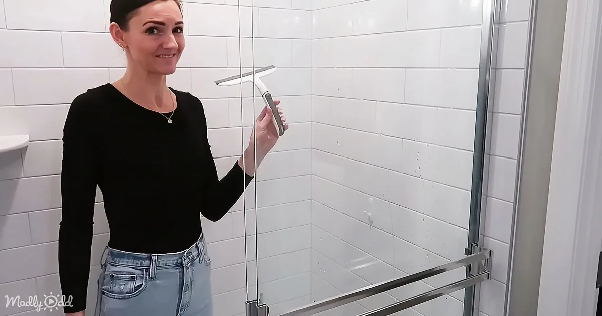 A woman in a black top and jeans holds a squeegee, smiling in a bathroom with white tiled walls and a glass shower door.