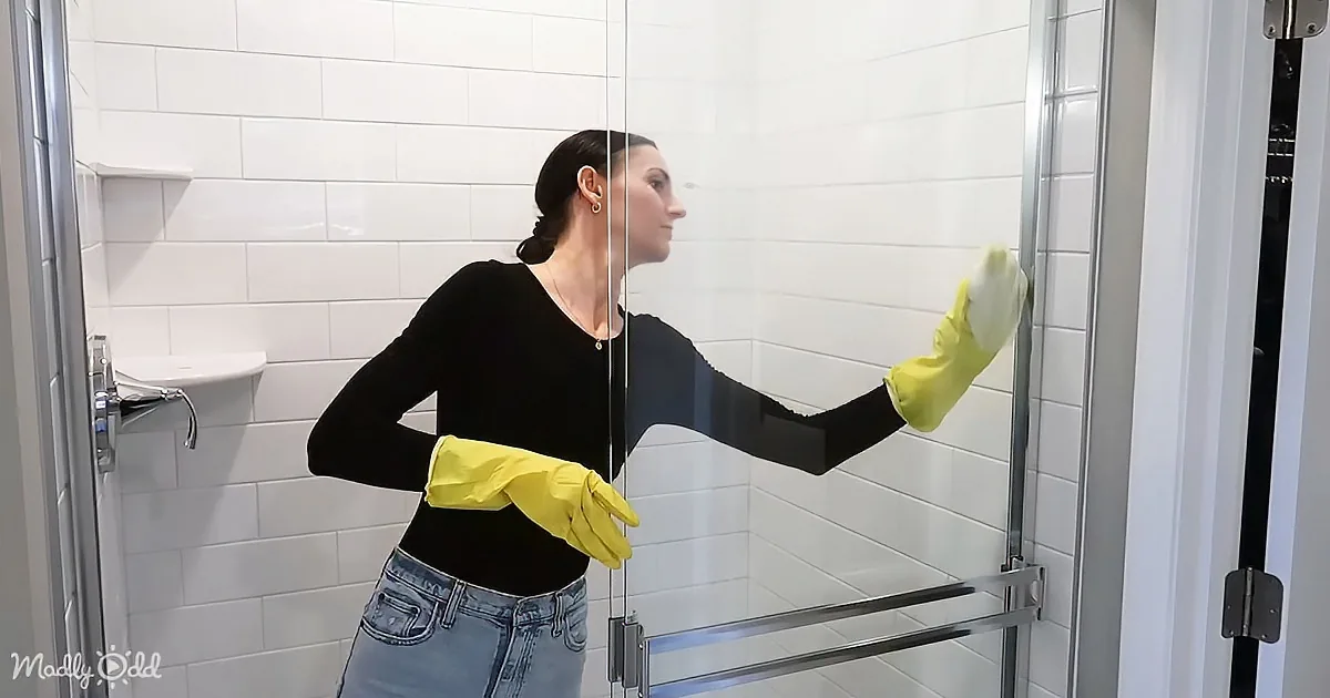 A woman in yellow gloves cleans a shower glass door with a white cloth.