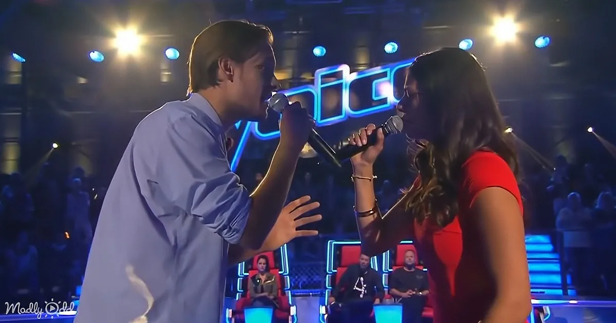 A man and woman sing into microphones on stage, with judges seated in red chairs in the background.