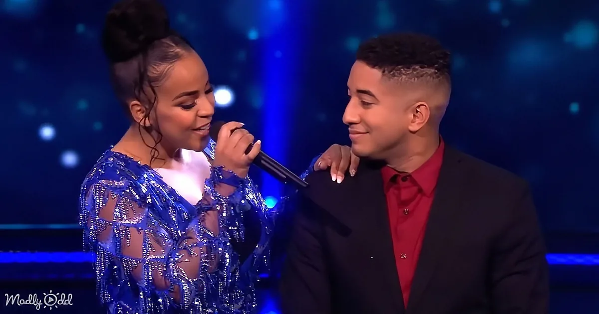 A woman in a sequined dress holds a microphone, smiling at a man in a suit with a red shirt, both on stage with blue lights.