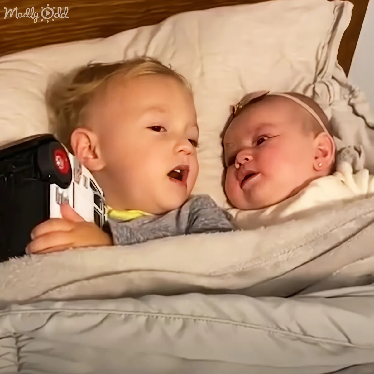A toddler and a baby are lying on a bed, the toddler holding a toy, both looking at each other.
