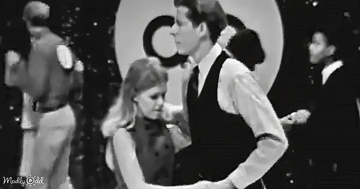 A couple in formal attire dances closely, surrounded by other dancers in a dimly lit setting.