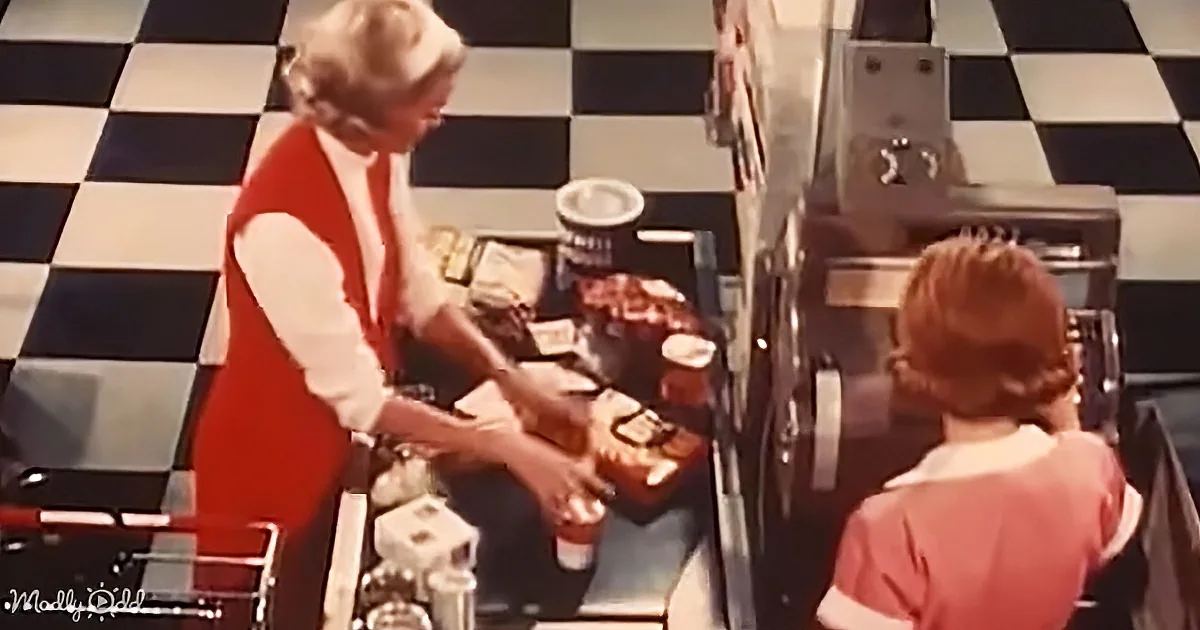 A woman in a red apron serves food at a counter with a checkered floor.