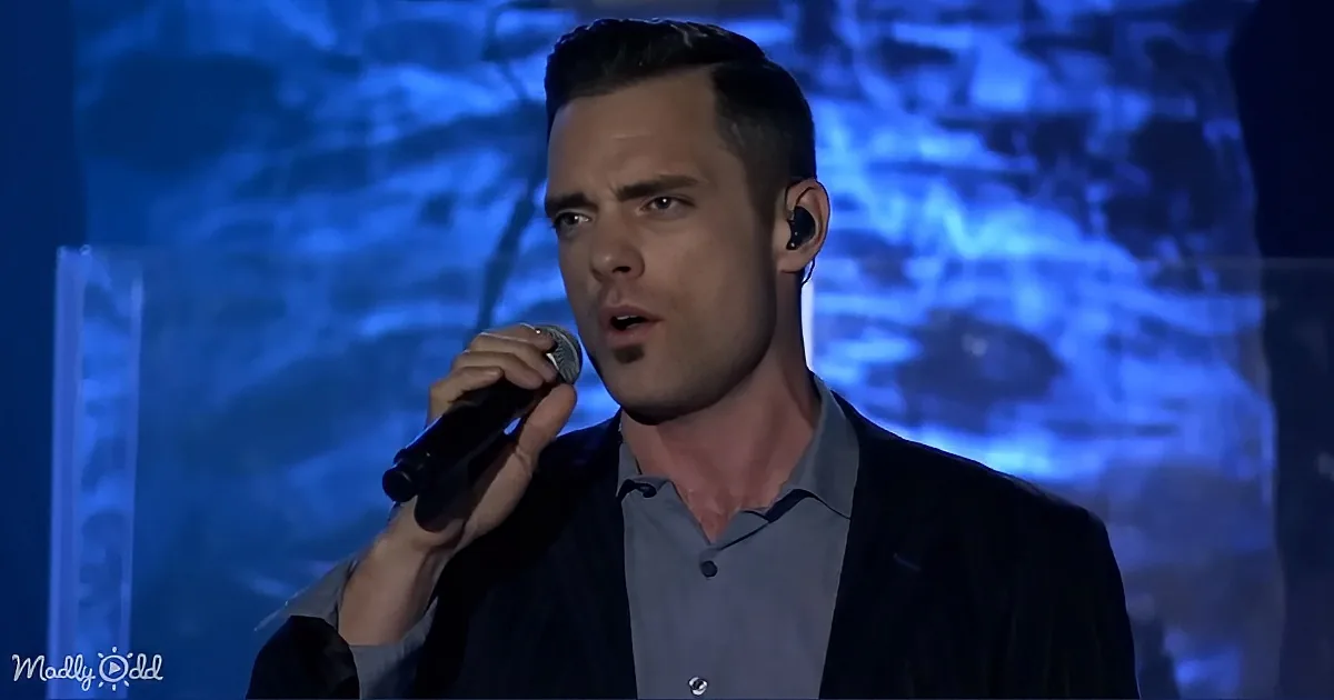 A man in a suit holds a microphone, singing on stage with a blue-lit background.