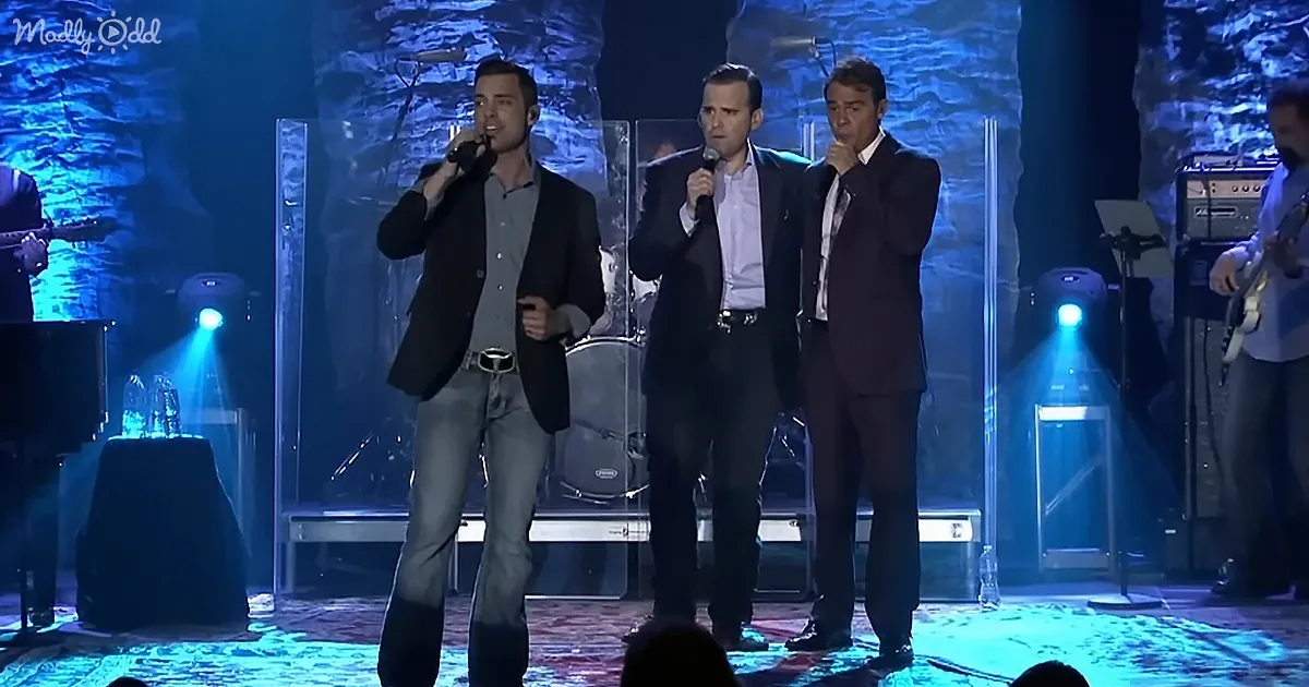 Three men in suits sing on stage with microphones, illuminated by blue lights.