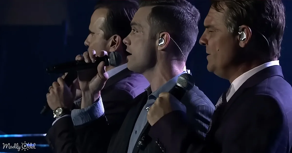 Three men in suits sing into microphones, wearing earpieces, in a dimly lit setting.