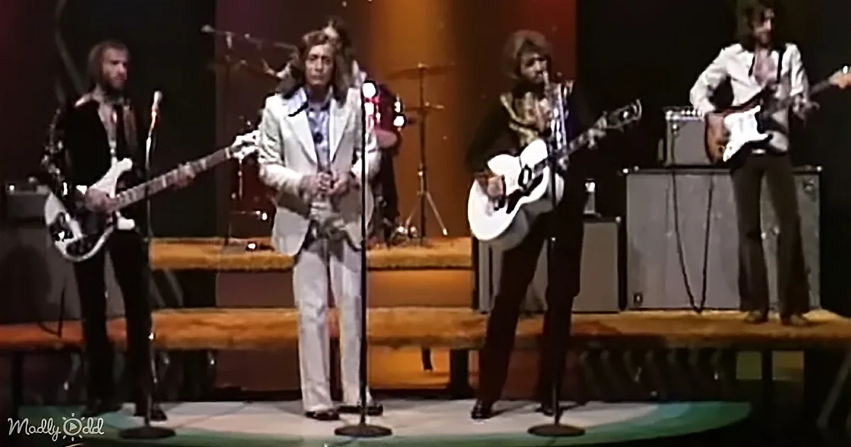 A band performs on stage with microphones, guitars, and a drum set, dressed in retro attire.