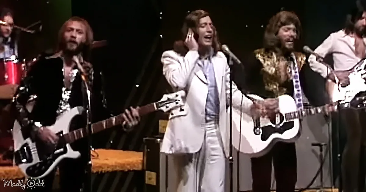 A man in a white suit sings passionately on stage with a band playing guitars.