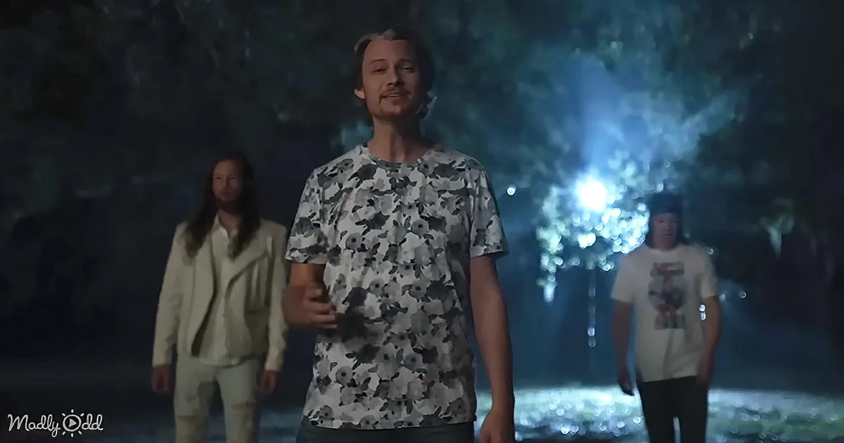 A man in a floral shirt stands in a dimly lit forest, with two others in the background, one in a white suit and another...