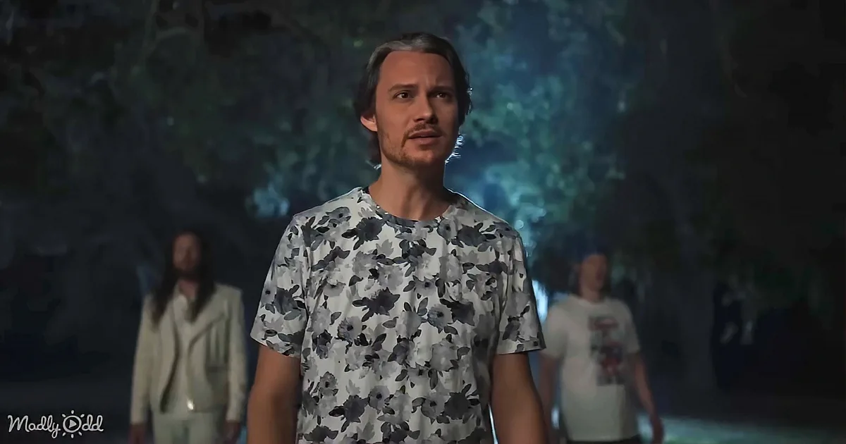 A man in a floral shirt stands in a dimly lit room, looking to the side with a concerned expression.