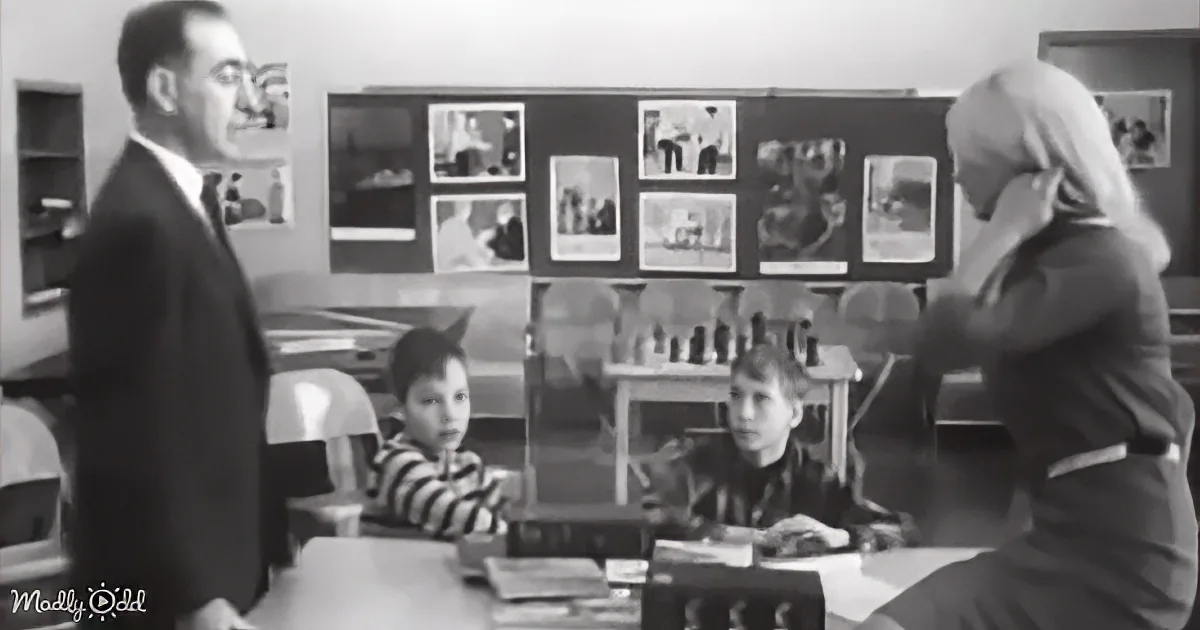 A teacher and a man in a suit stand in front of two seated students in a classroom.
