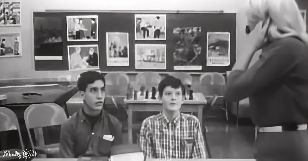 A teacher stands at the front, addressing students in a classroom with a bulletin board of photos in the background.