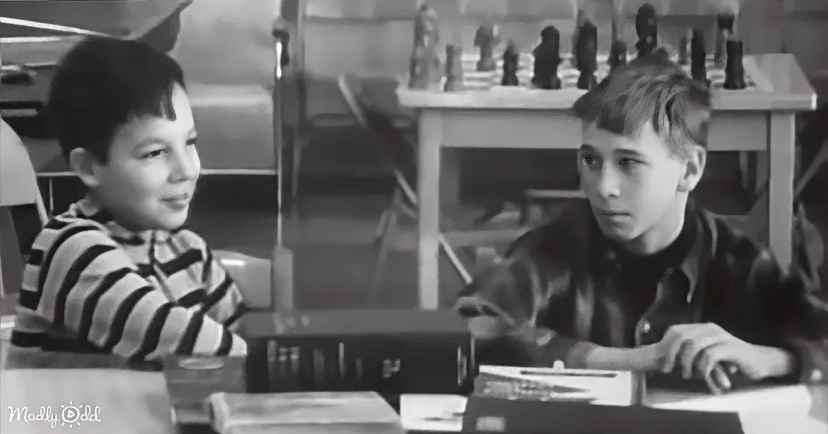 Two boys sit at a table, one in a striped shirt, the other in a dark shirt, with a chessboard in the background.