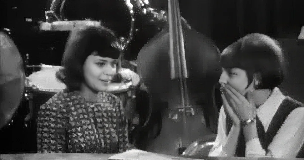 Two women, one with a cigarette, sit at a table, engaged in a conversation, with a cello in the background.